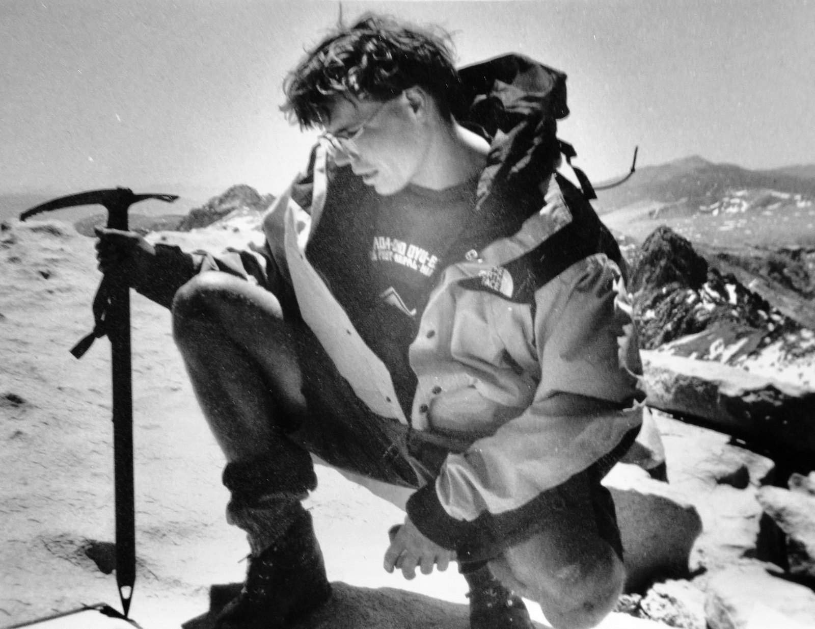 Black and white photograph of Brooks crouched on the summit of Mt. Whitney reading the elevation marker, ice axe planted beside him