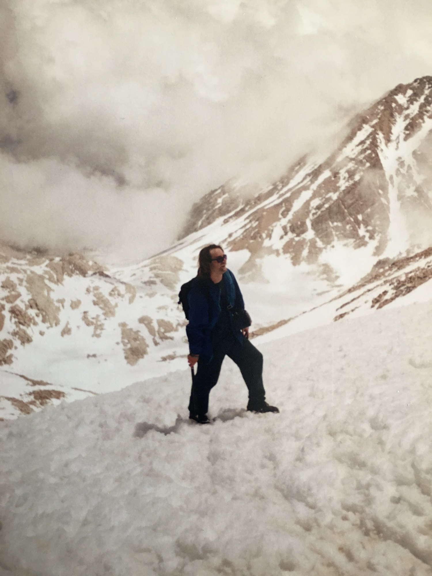 Zack Hurley standing on a snow slope below the Whitney crest, ice axe in hand, cloud pouring over the ridge behind him.